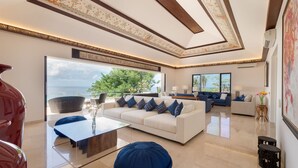 A spacious living room with a white sofa, blue cushions, a glass coffee table, and a blue ottoman. There is a large window with a view of the ocean. - Pandawa Cliff Estate (Kutuh)