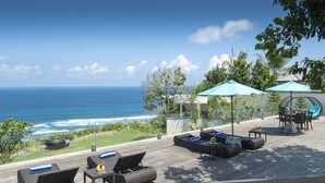 A wooden deck with outdoor furniture, including sofas and a table, overlooking a beach with the ocean in the background. - Pandawa Cliff Estate (Kutuh)