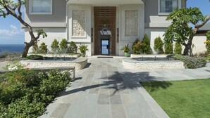 A two-story house with a stone pathway leading to a wooden door, surrounded by landscaped gardens and trees. - Pandawa Cliff Estate (Kutuh)