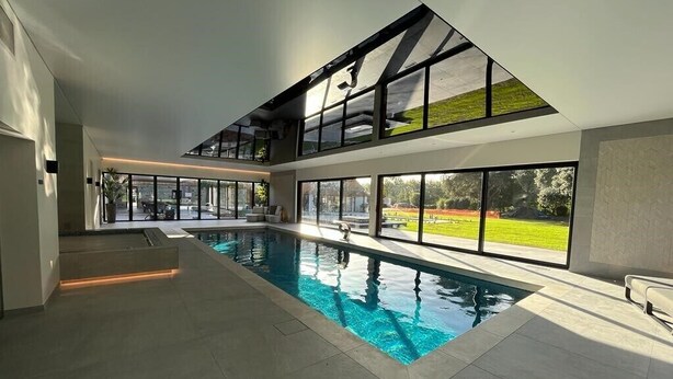 Una piscina interior amplia con diseño moderno, grandes ventanales que ofrecen vistas a un jardín paisajístico y un techo alto con tragaluces.