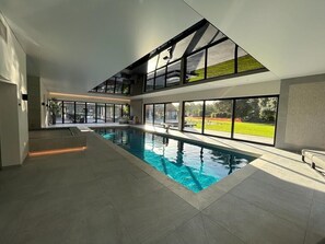 Een ruime overdekte zwembad met een modern design, grote ramen met uitzicht op een aangelegde tuin en een hoge plafond met dakramen.