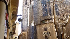 Une rue étroite avec une terrasse de café dotée de sièges en plein air et de parasols, un bâtiment historique avec des fenêtres en arc de cercle, et un arbre au premier plan.