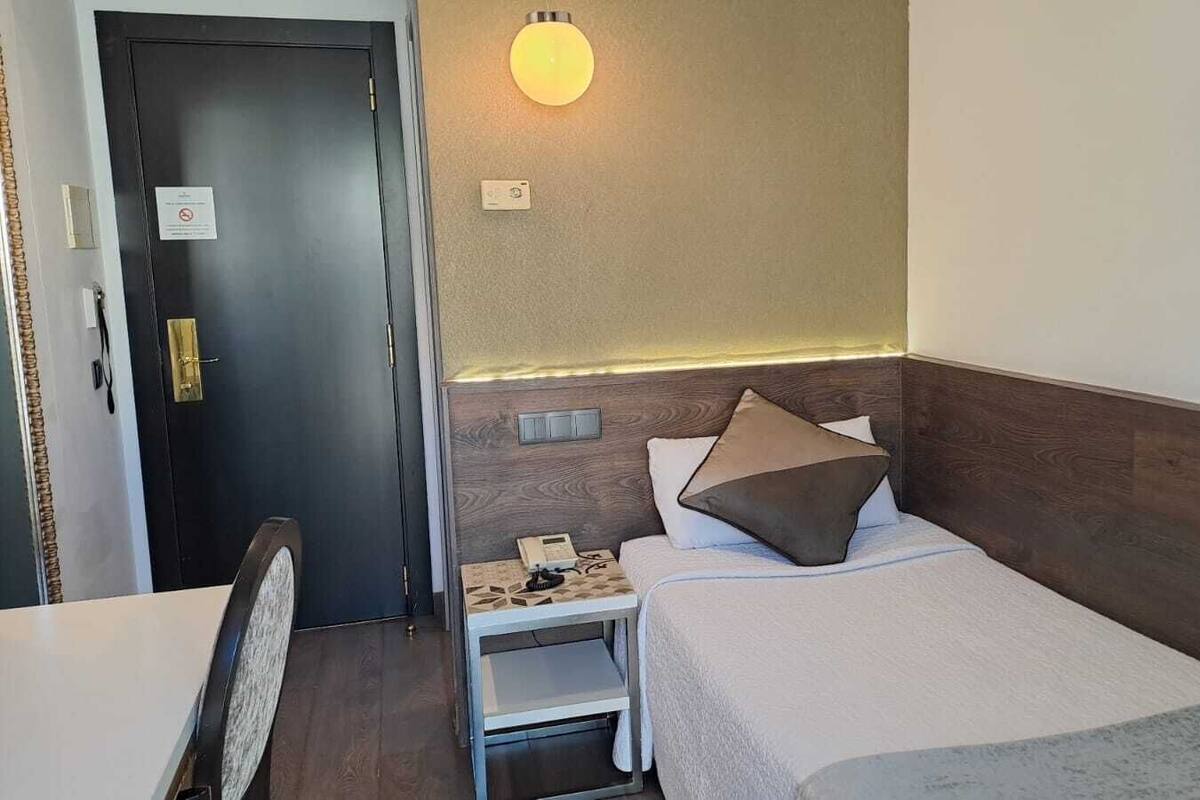 A hotel room with a bed, bedside table, lamp, and a door.
