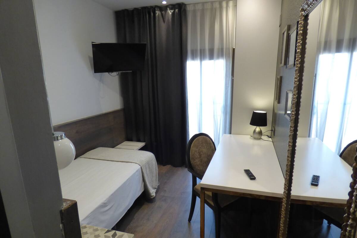 A hotel room with a bed, a desk, a chair, a TV mounted on the wall, and a mirror.