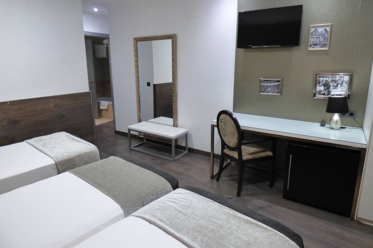 A hotel room with two single beds, a desk with a chair, a mirror, a TV, and a small refrigerator.
