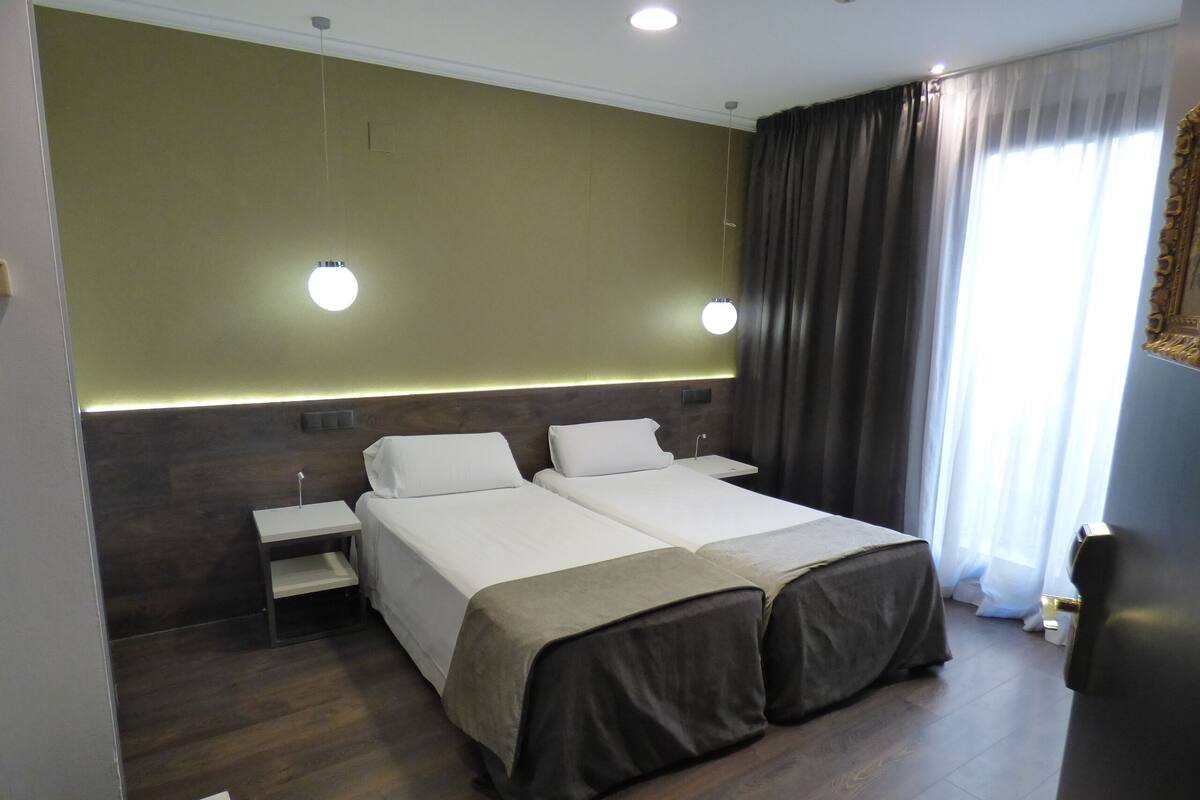 A hotel room with two beds, a nightstand, a lamp, and a window with curtains.