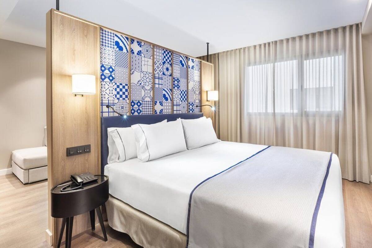 Hotellihuone, jossa on suuri sänky, yöpöytä, jossa on puhelin, koristeellinen sängynpääty ja ikkuna, jossa on verhot.