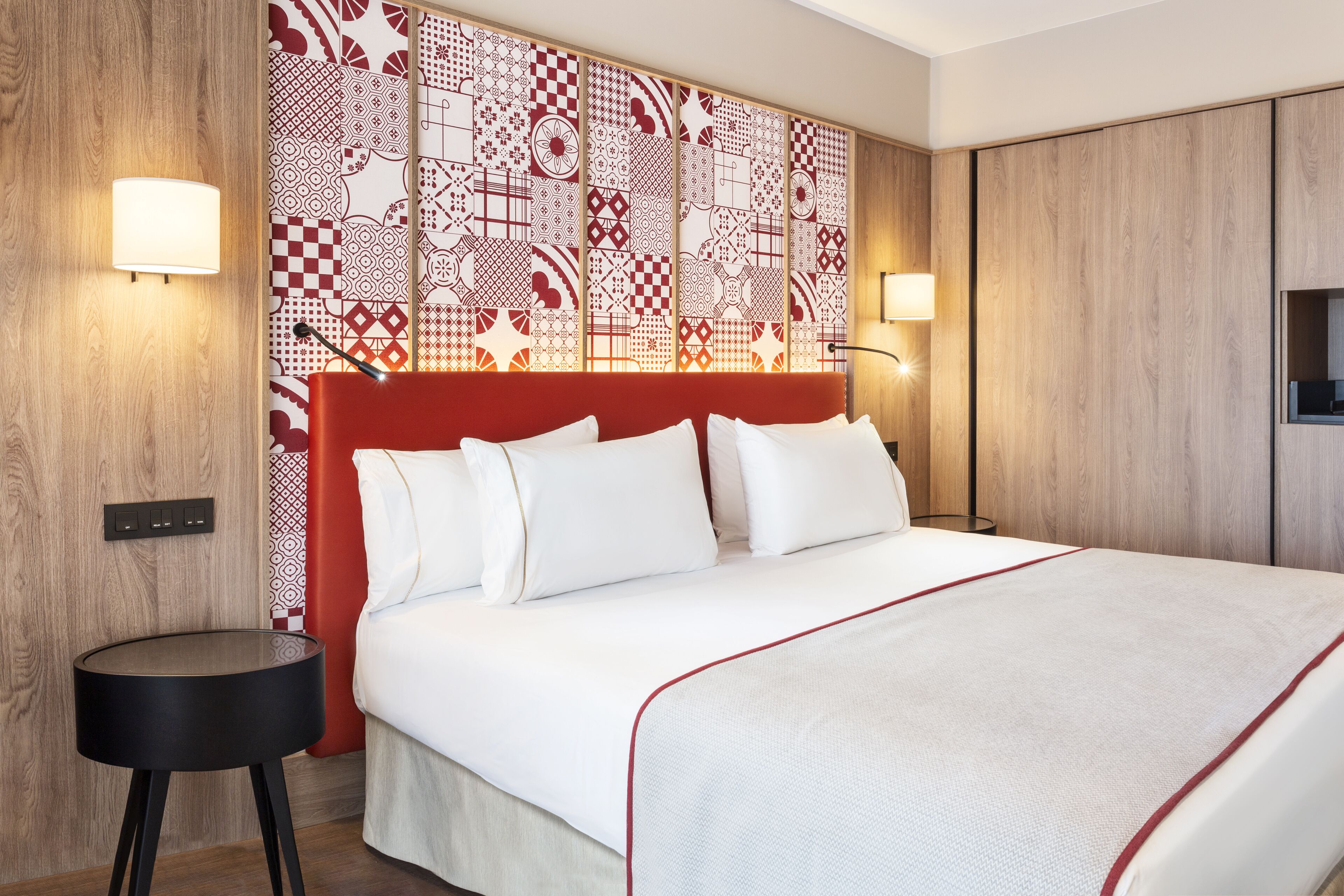 A modern hotel room with a large bed, a red headboard, and patterned wallpaper. There are two bedside lamps and a small black side table.