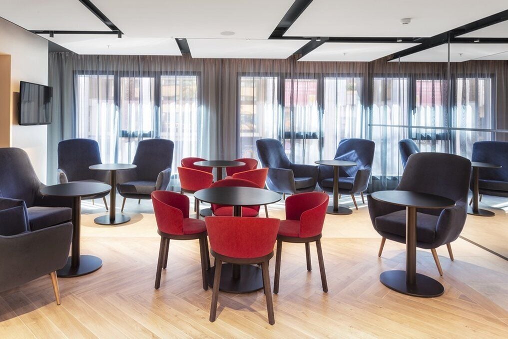 Un moderno vestíbulo de hotel con una mezcla de sillas negras y rojas alrededor de mesas, ventanas grandes con cortinas y un televisor de pantalla plana montado en la pared.