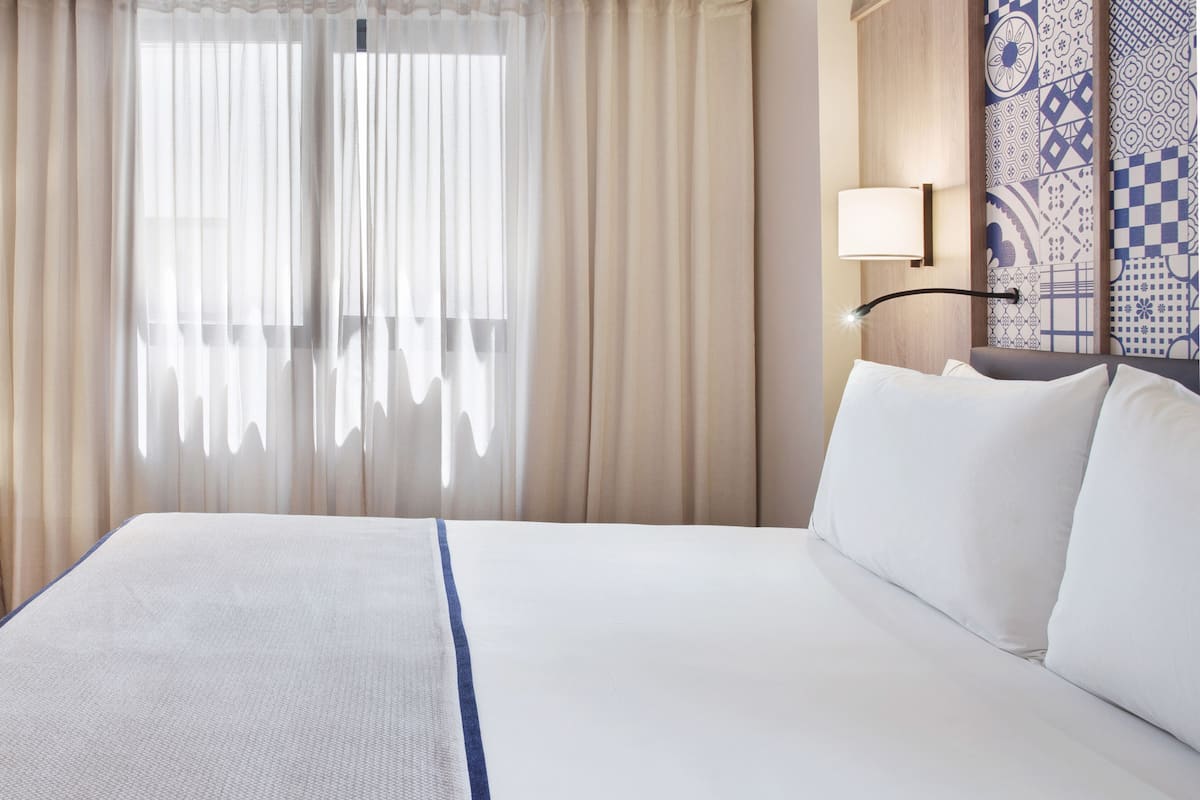 Hotellihuone, jossa on suuri sänky, läpikuultavat verhot, seinään kiinnitetty lamppu ja kuviollinen tapetti.