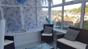 A sunlit room with wicker furniture, a glass coffee table, and a view of a residential area through large windows. - Tranquil Retreat - 3 Bedroom House With hot tub (Torquay)