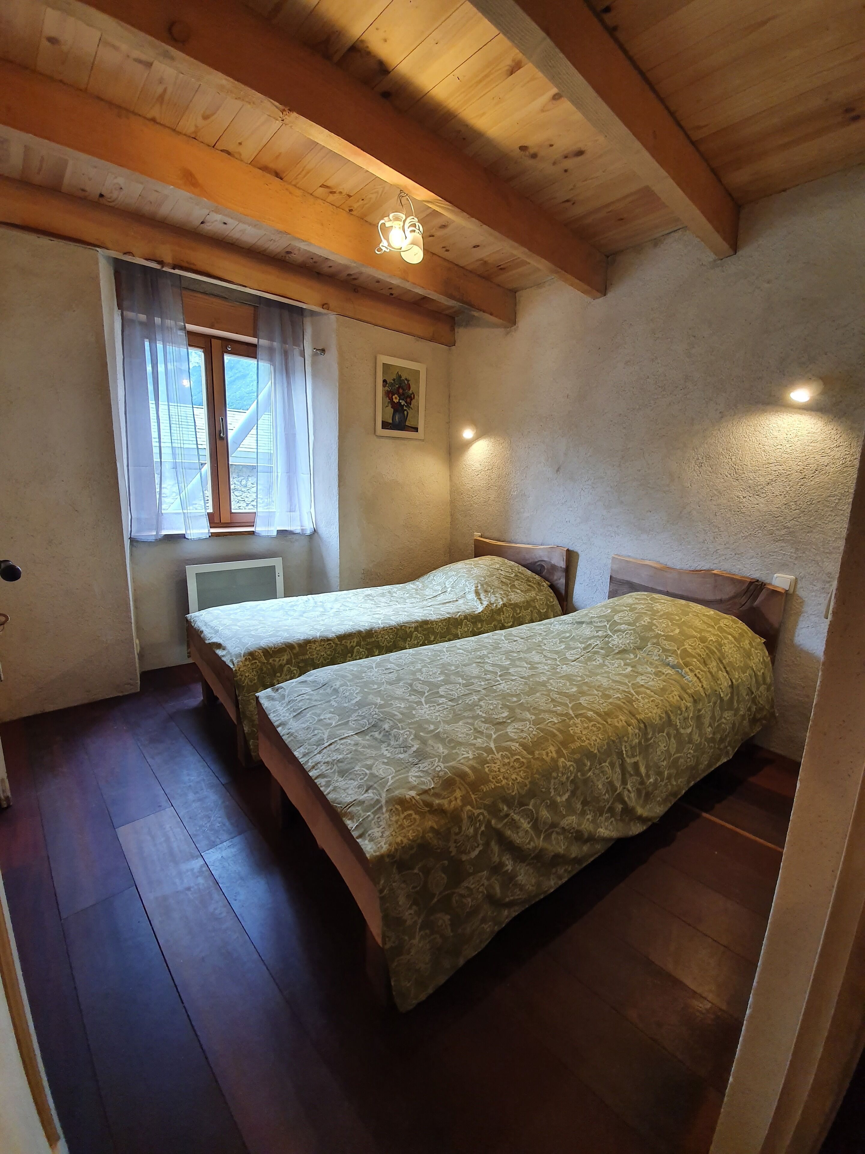 Une petite pièce accueillante avec deux lits, des poutres en bois au plafond, une fenêtre ornée de rideaux légers et un tableau accroché au mur.
