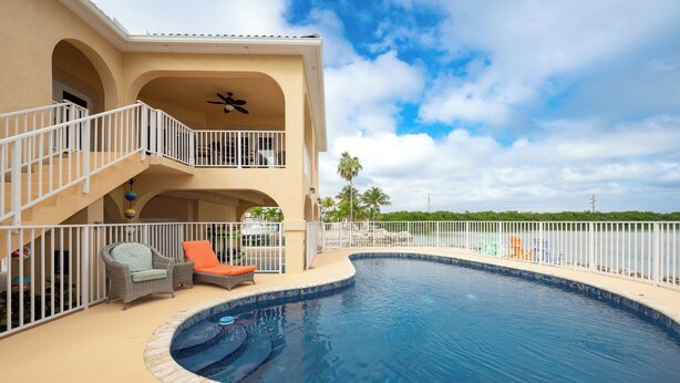 A swimming pool with a curved edge, a covered seating area with chairs, and a balcony with railings. - Captains Quarters - Cudjoe Key - Pool- Waterfront w/ Boat Dock (Cudjoe Key)
