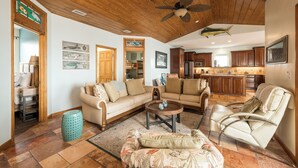 A spacious living room with a wooden ceiling, a ceiling fan, a large sofa, a coffee table, and a dining area with wooden cabinets and a kitchen island. - Captains Quarters - Cudjoe Key - Pool- Waterfront w/ Boat Dock (Cudjoe Key)