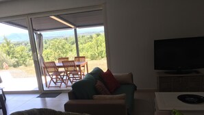 Ein Wohnzimmer mit Sofa, Sessel, Fernseher und Blick auf eine Terrasse mit Tisch und Stühlen.