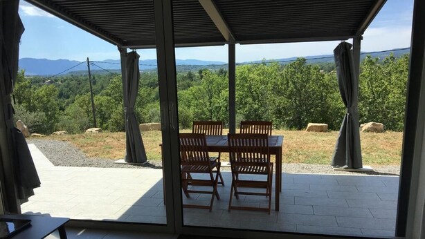 Eine überdachte Terrasse mit Tisch und Stühlen, Blick auf eine gebirgige Landschaft und eine Steinmauer.