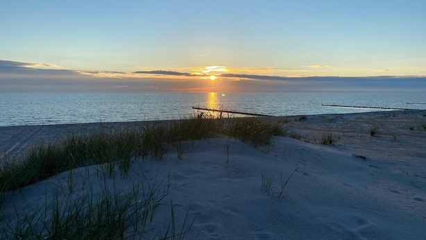 Ein Strand mit Sonnenuntergang über dem Wasser, Dünen und vereinzelter Vegetation.