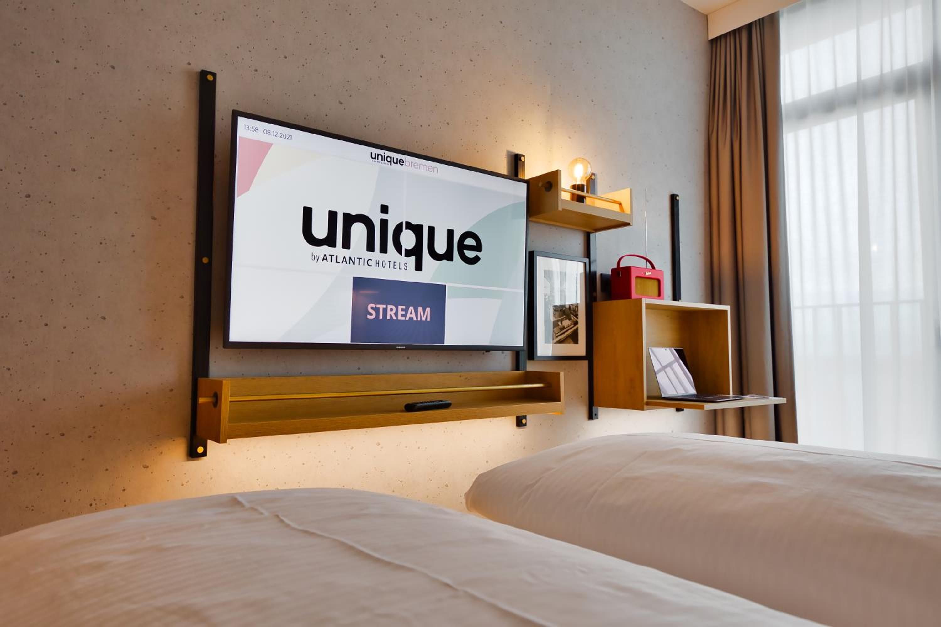 Quarto de hotel com televisão fixada na parede exibindo o logotipo 'unique by ATLANTIC HOTELS', uma estante de madeira com abajur e uma cama com roupa de cama branca.