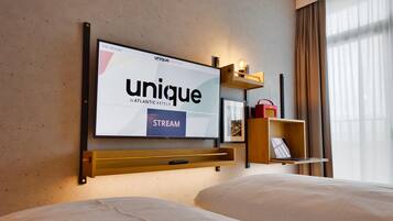 Quarto de hotel com televisão fixada na parede exibindo o logotipo 'unique by ATLANTIC HOTELS', uma estante de madeira com abajur e uma cama com roupa de cama branca.