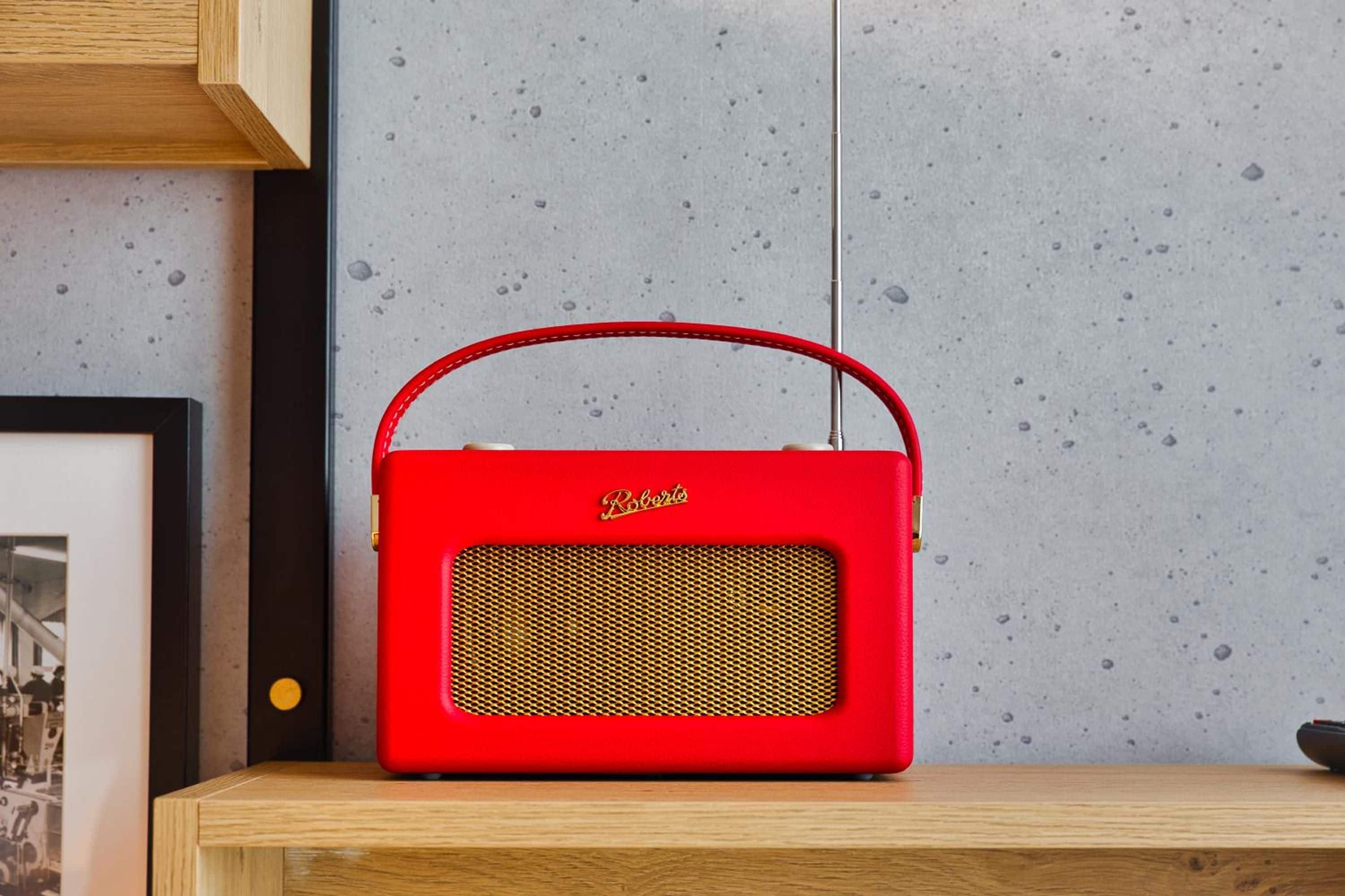 A red portable speaker with a handle on a wooden surface against a speckled wall.