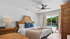 Un dormitorio con cama, mesita de noche de madera, mesa, ventilador de techo y vista a una piscina a través de una puerta corrediza de vidrio.