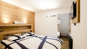 A bedroom with a wooden headboard, a bed with a striped bedspread, a bedside table with a lamp, and a television mounted on the wall. - Apartment Véronique, front of village location (Montgenevre)