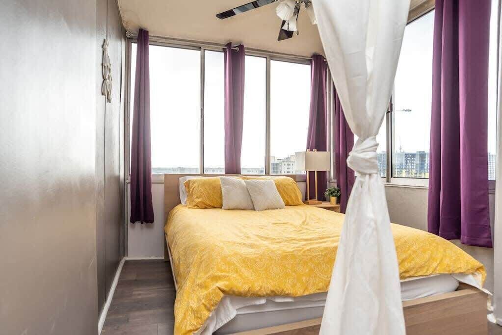 A bedroom with a bed, curtains, a ceiling fan, and a view of the city.