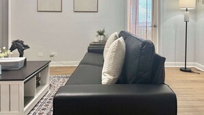 A modern living room with a black leather sofa, two framed artworks, a small table with decorative items, a floor lamp, and a door with a view. - Condo in the Heart Atlanta w/ Private Balcony (Atlanta)