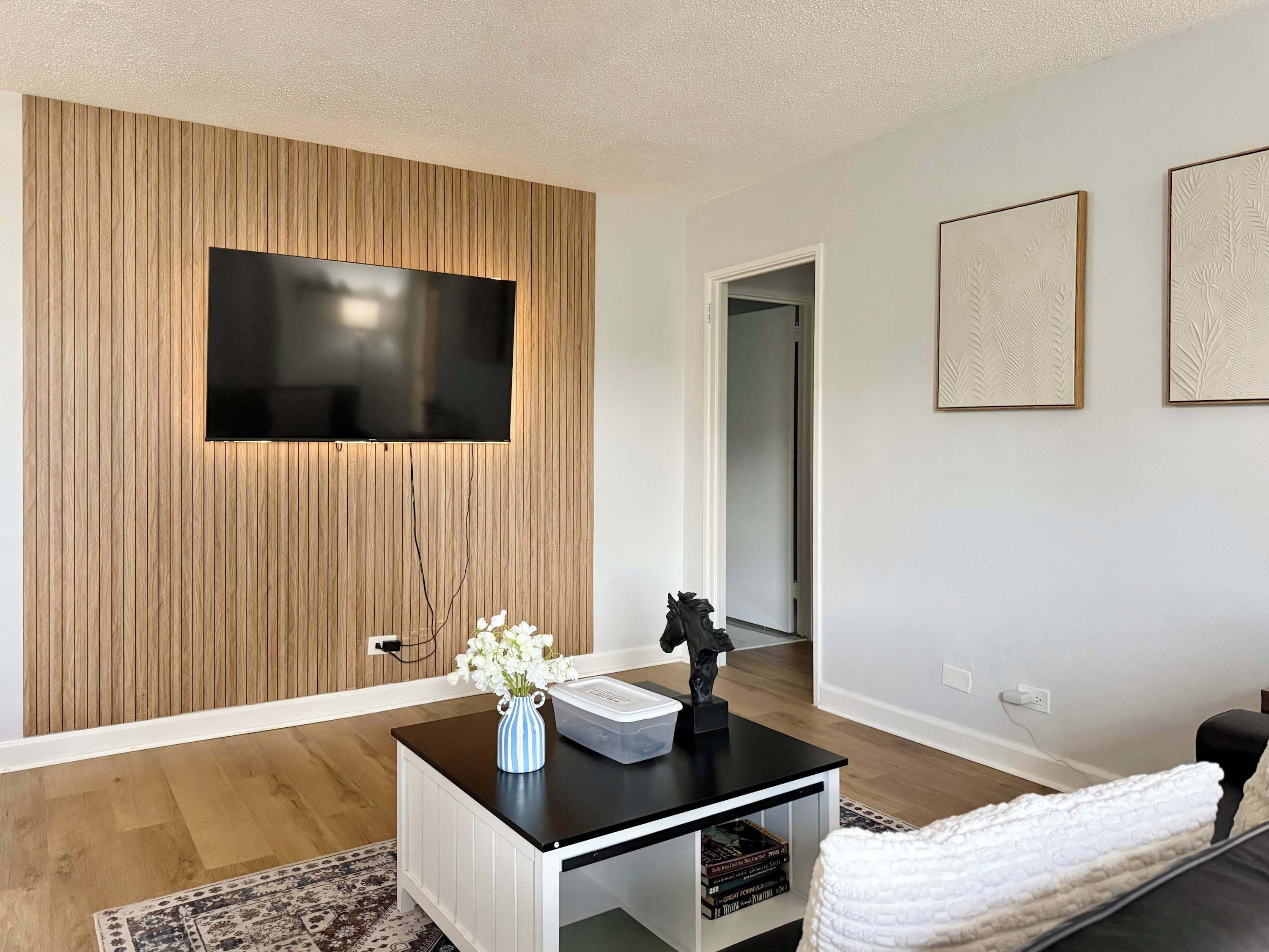 A modern living room with a flat-screen TV mounted on a wooden panel wall, a coffee table with a vase of flowers, a statue, and a sofa.