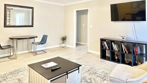 Una sala de estar con televisor, mesa de comedor, sillas, sofá, mesa de centro, estantería y una planta.