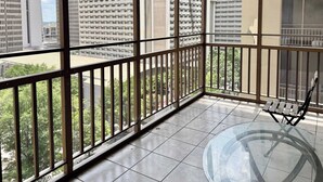 Un balcón con mesa de vidrio y barandilla metálica, con vistas a un paisaje urbano de edificios altos.