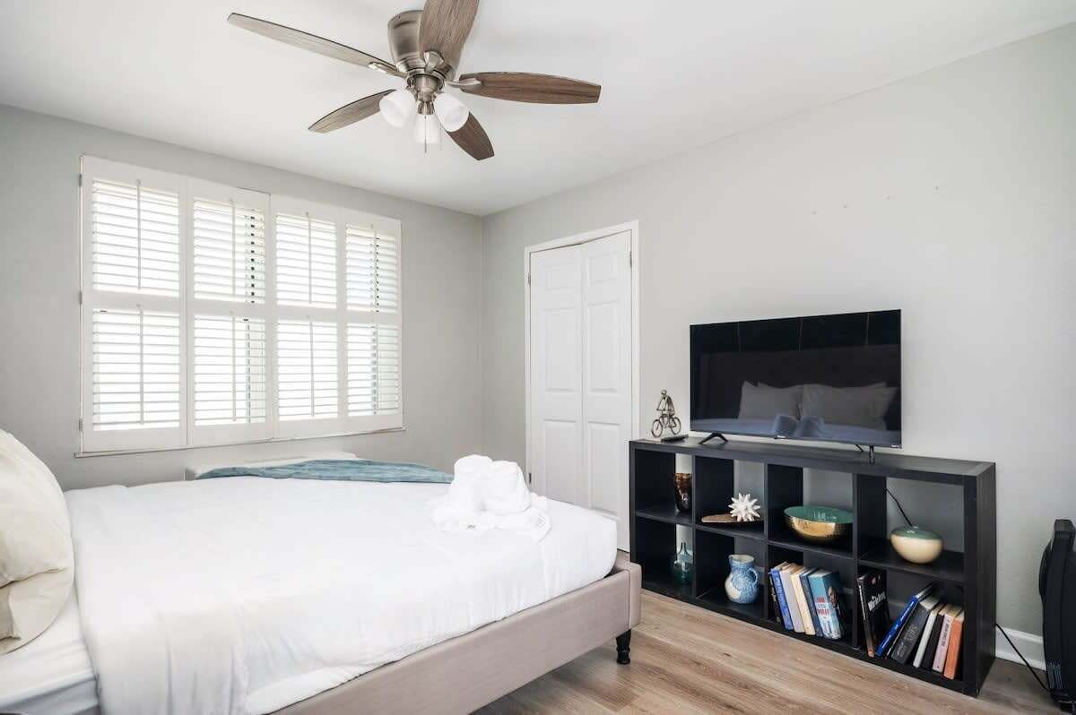 A bedroom with a bed, a ceiling fan, a TV on a black stand, and a bookshelf with books and decorative items.