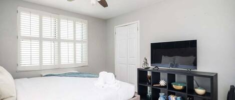 A bedroom with a bed, a ceiling fan, a TV on a black stand, and a bookshelf with books and decorative items.