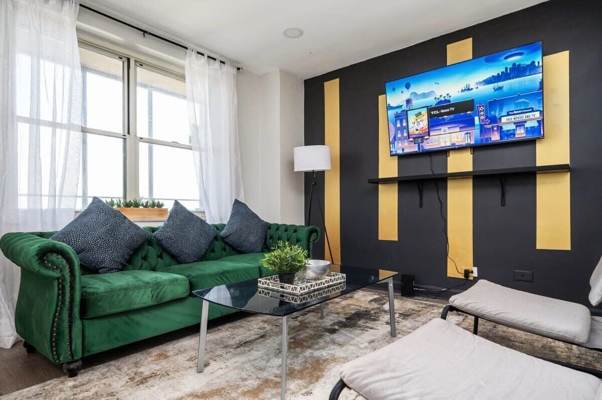 Un salon moderne avec un canapé en velours vert, une table basse en verre et un téléviseur à écran plat fixé au mur.
