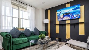 A modern living room with a green velvet sofa, a glass coffee table, and a flat-screen TV mounted on the wall.