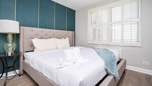 A neatly made bed with white linens and a blue throw blanket, a bedside table with a lamp, and a window with white shutters.