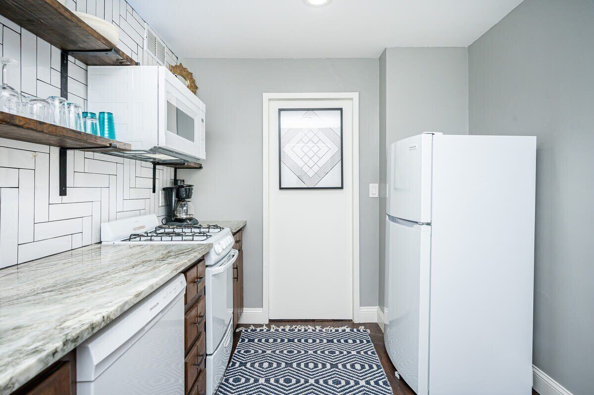 Cocina compacta con armarios blancos, estufa a gas, refrigerador blanco y una alfombra estampada.