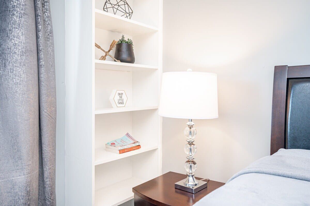 Une chambre d’hôtel bien rangée, avec un lit, une table de chevet dotée d’une lampe, une étagère ornée d’objets décoratifs et une fenêtre avec des rideaux.