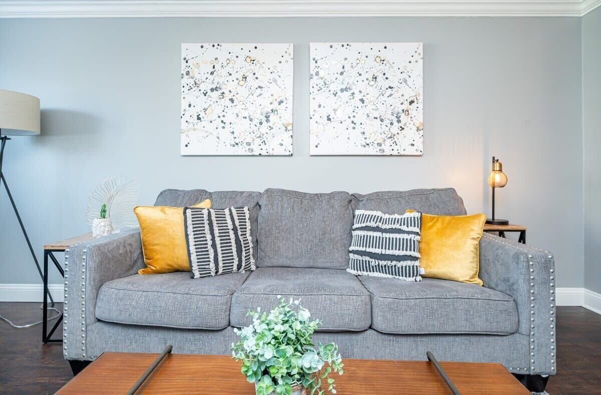 Ein graues, gepolstertes Sofa mit Zierkissen, ein Couchtisch aus Holz mit einer Pflanze und zwei abstrakte Gemälde an der Wand.
