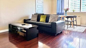 A living room with a black leather sofa, a glass coffee table, a dining area with a table and chairs, and a window with a view of buildings. - Upgraded 2BR Condo Near ATL Attractions (Atlanta)