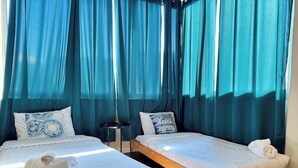 Una habitación con dos camas, cada una con un colchón blanco y almohadas a juego, una mesita redonda pequeña y una ventana con cortinas azules.
