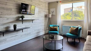 Un moderno salón con una televisión de pantalla plana, un ventilador de techo y un amplio ventanal con vistas a edificios y árboles.
