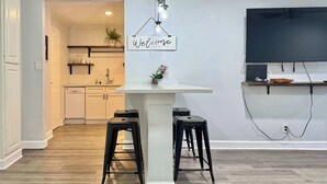 A modern kitchen with a central island, black bar stools, a 'Welcome to our place' sign, a TV mounted on the wall, and a wooden floor. - Downtown Atlanta Condo with City View (Atlanta)