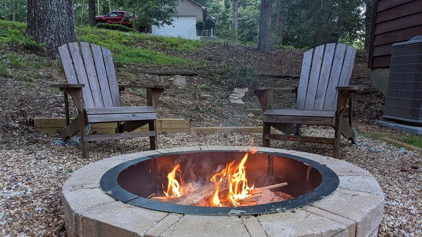 Un foyer circulaire, avec des flammes, entouré d’une bordure en pierre, et deux fauteuils Adirondack placés face à face.