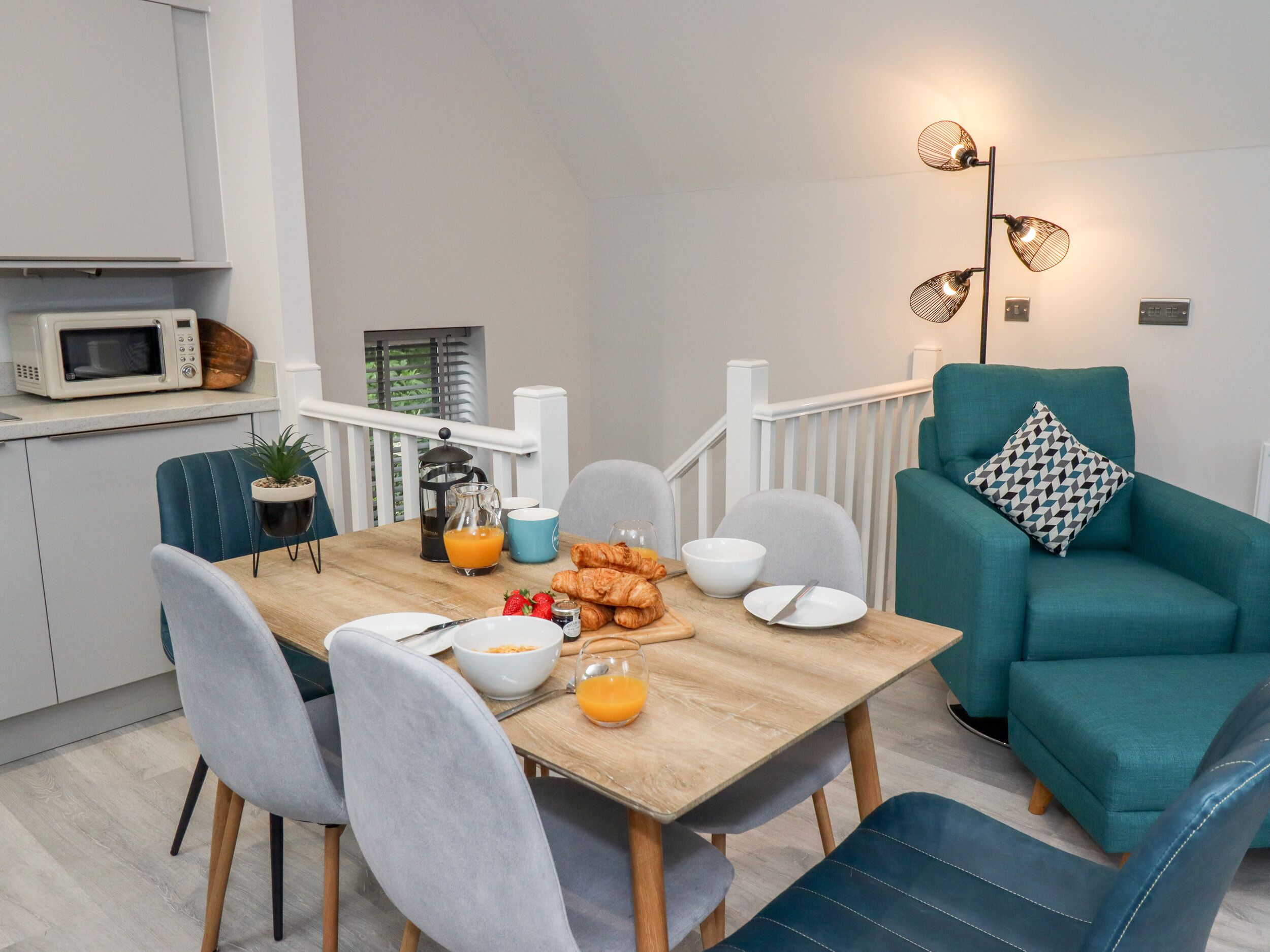 Una cocina moderna con microondas, cafetera y frutero. Hay una mesa de comedor con sillas y un sofá color turquesa con un cojín estampado.