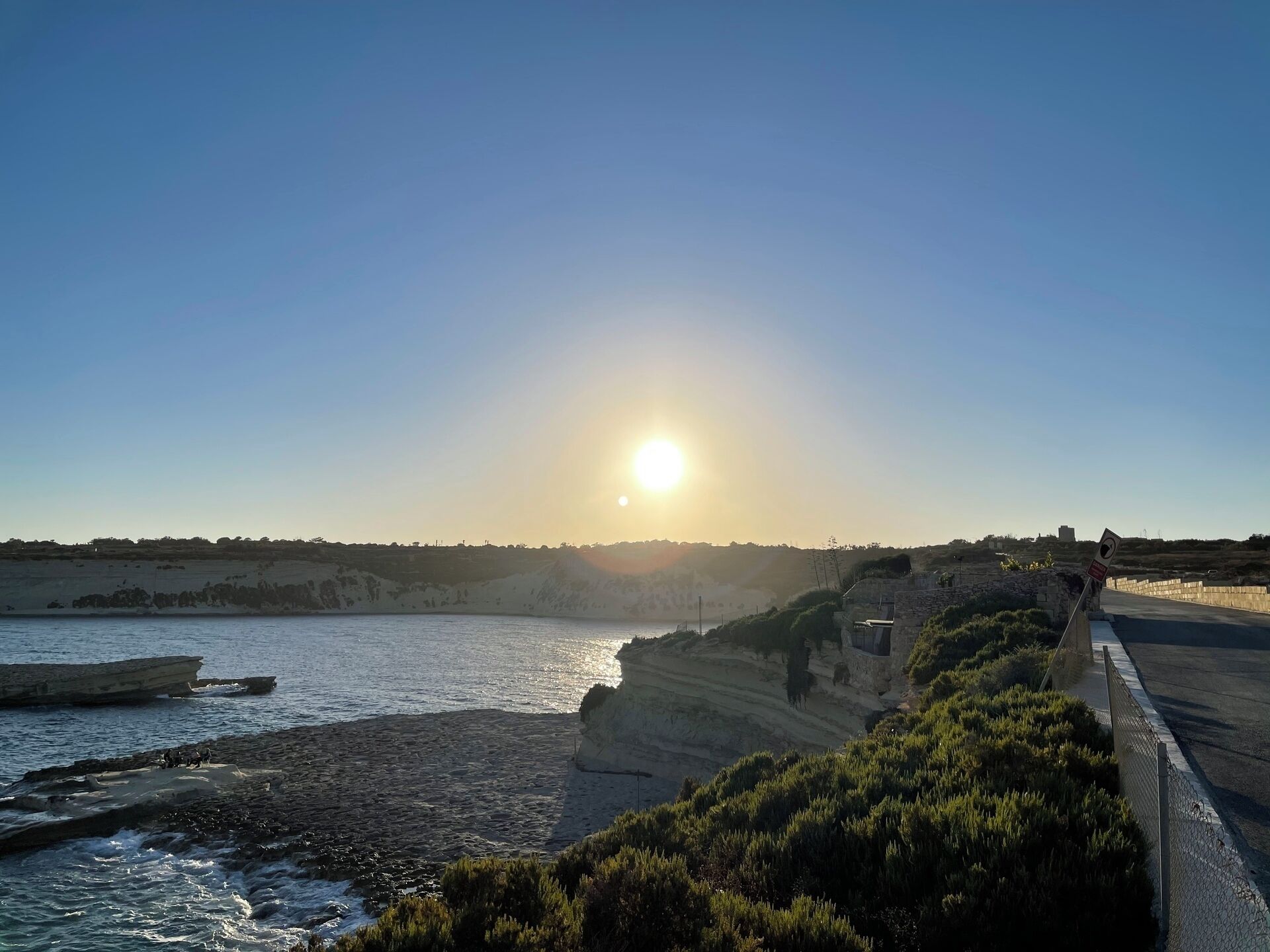 A coastal landscape with a clear sky, a sun low on the horizon, and a stone wall running parallel to the water.