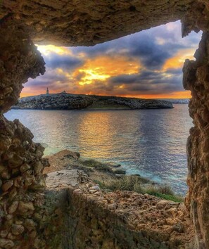 A coastal view through a rocky archway with a lighthouse in the distance. - Sunset Modern Apt - Near Beach, Wifi & Cable TV (Birzebbuga)