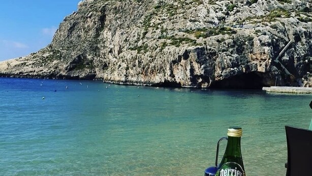 Una mesa con un plato de comida, una botella de agua Perrier y una cerveza, frente a un pintoresco paisaje costero.