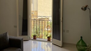 Una habitación con un sofá gris, un jarrón verde y una puerta corredera abierta que da a un balcón con plantas en macetas.