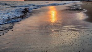 Una playa al atardecer, con el sol bajo en el horizonte, olas suaves lamiendo la orilla y una montaña al fondo.